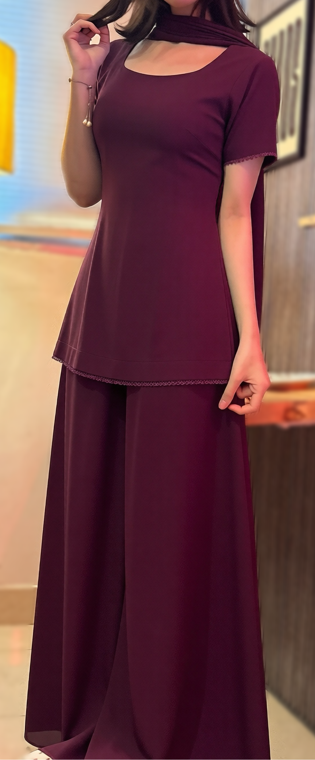 A woman modeling a matching two-piece outfit consisting of a short-sleeve scoop-neck top and wide-leg trousers, both in a deep plum or burgundy shade. She is standing in an indoor room with a floor lamp, wall decor, and shelves in the background.