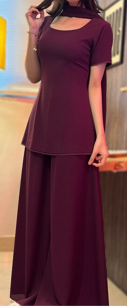 A woman modeling a matching two-piece outfit consisting of a short-sleeve scoop-neck top and wide-leg trousers, both in a deep plum or burgundy shade. She is standing in an indoor room with a floor lamp, wall decor, and shelves in the background.