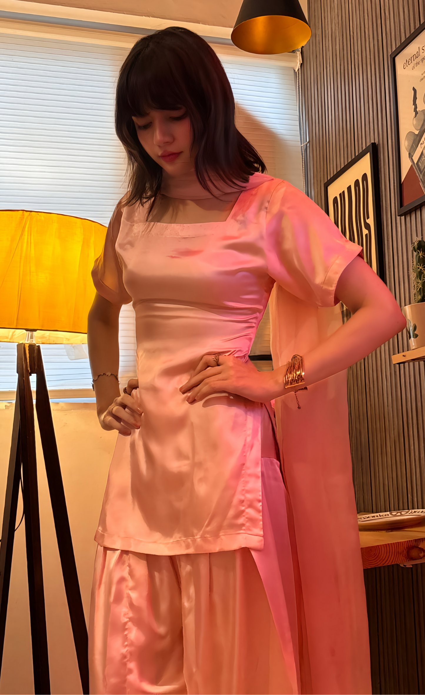 A woman wearing a light pink satin kurta and matching trousers with a sheer pink dupatta, standing in a warmly lit room with a floor lamp, window blinds, and framed wall art.