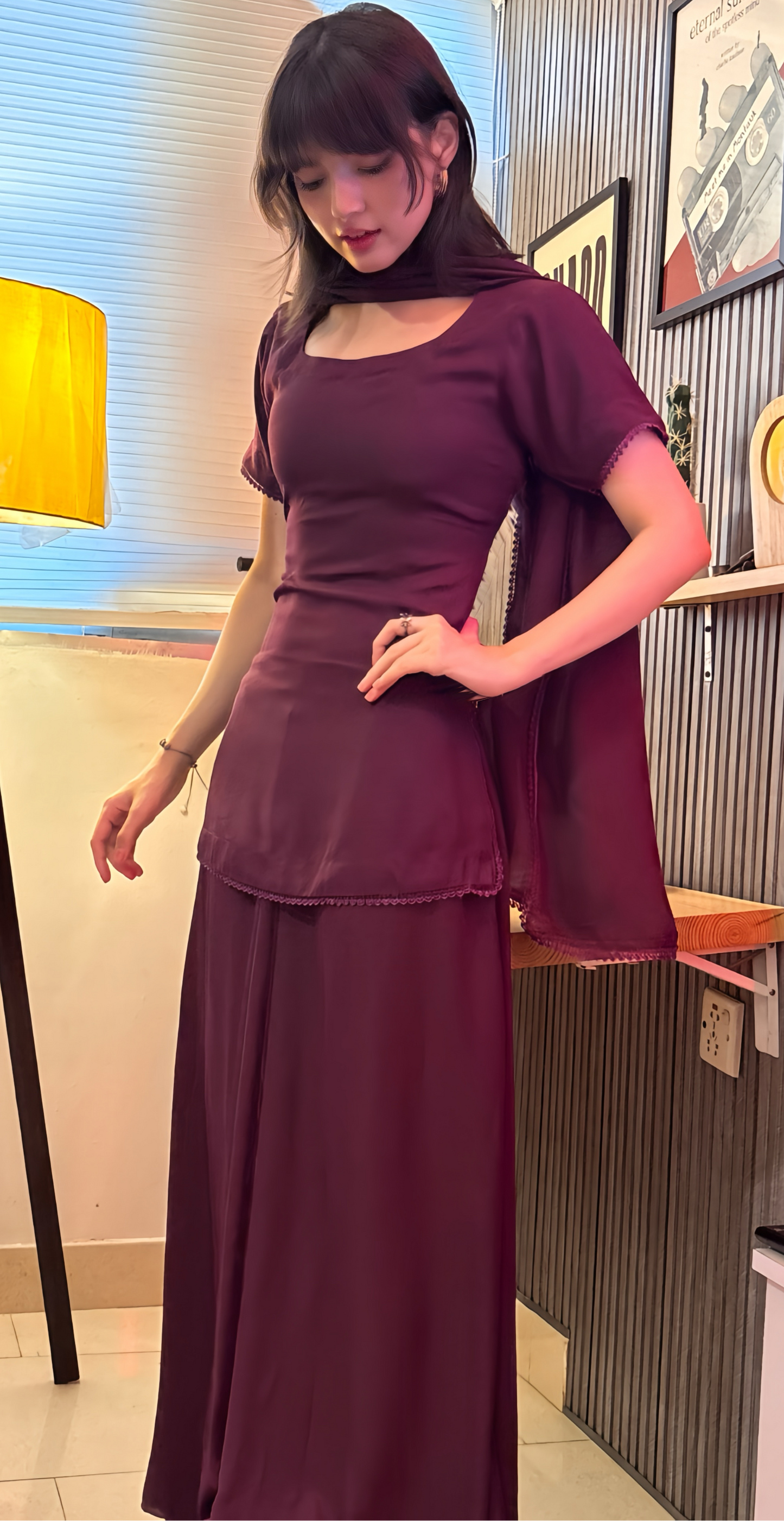 A woman models a dark plum purple two-piece outfit consisting of a short-sleeve top with a square neckline and a matching long skirt, standing in an indoor room with a floor lamp and wall decor.